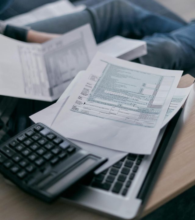 A young adult using a calculator and laptop to manage finances at home, surrounded by papers.