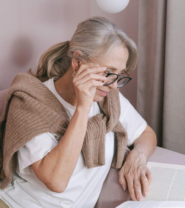 Elderly woman with glasses reading on a couch for leisure at home.